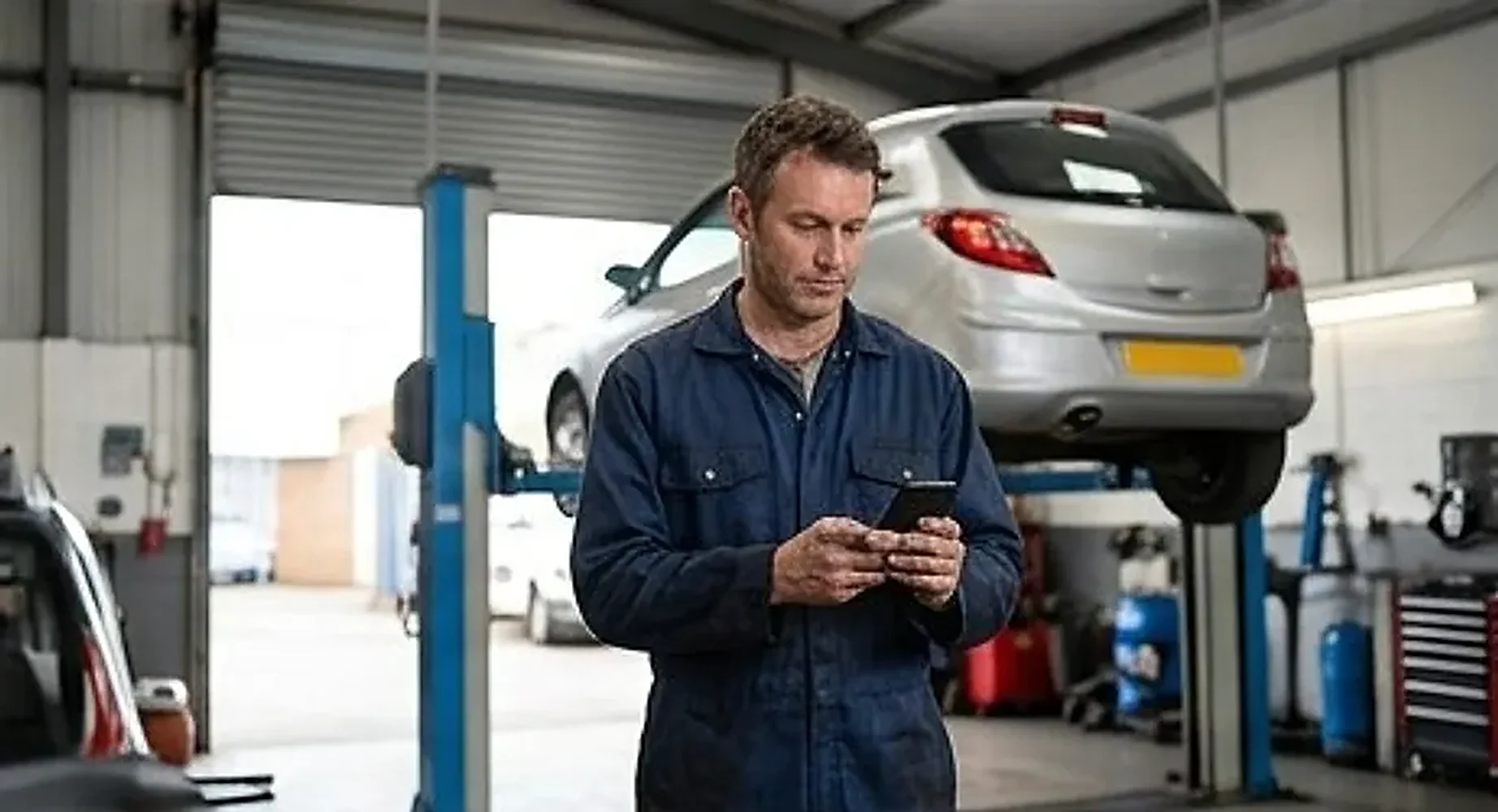 Mechanic in a garage bay reading a message with a car raised on the ramp behind him
